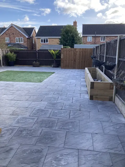 Grey riven slab patio with a timber sleeper planter and a patch of artificial grass